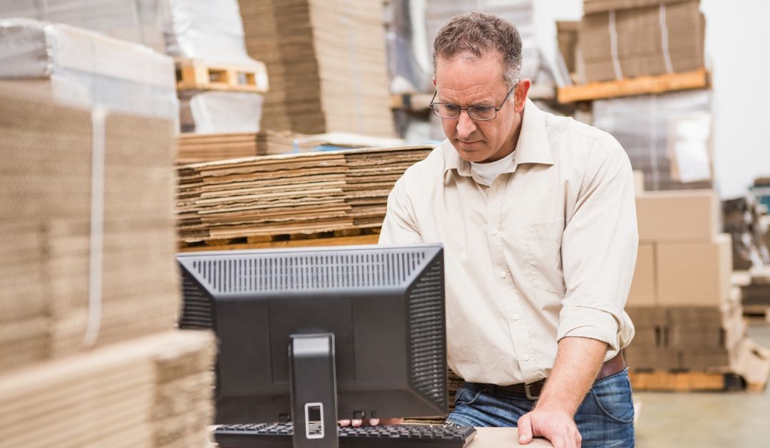 A man within a warehouse looks with a focus on a computer in front of him. He is surrounded by multiple stacks of flat boxes.