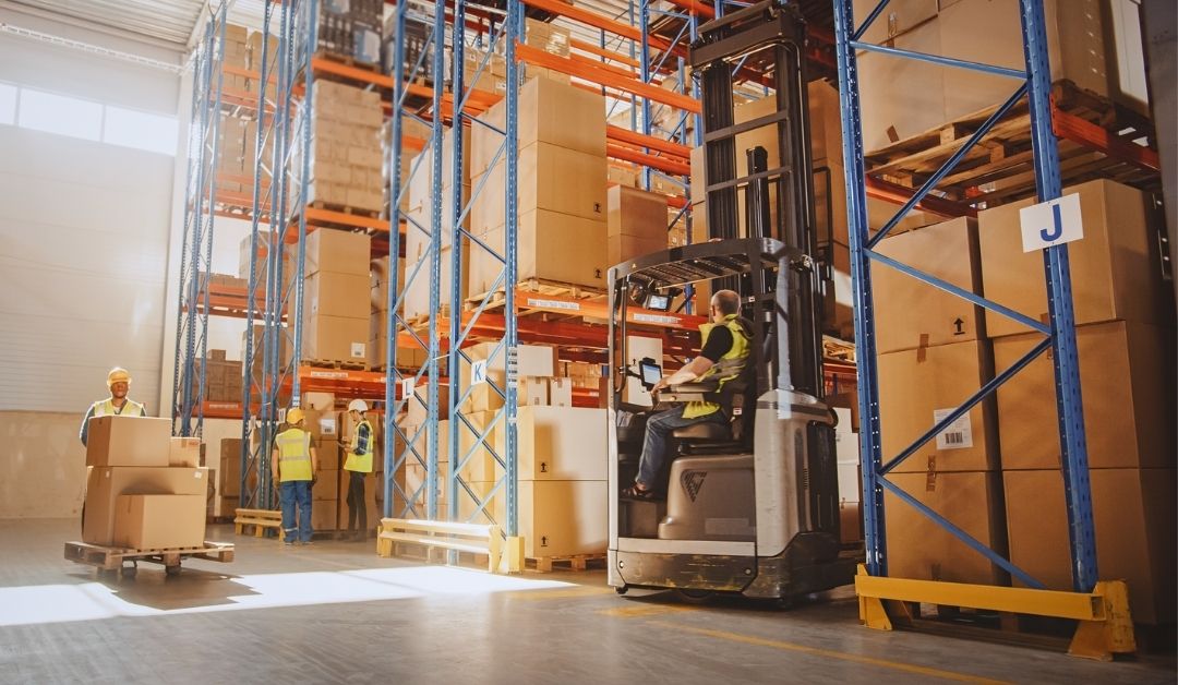 A small group of warehouse workers are completing various warehouse tasks such as driving a forklift and taking inventory. They are surrounded by floor to ceiling warehouse shelves stacked with large boxes