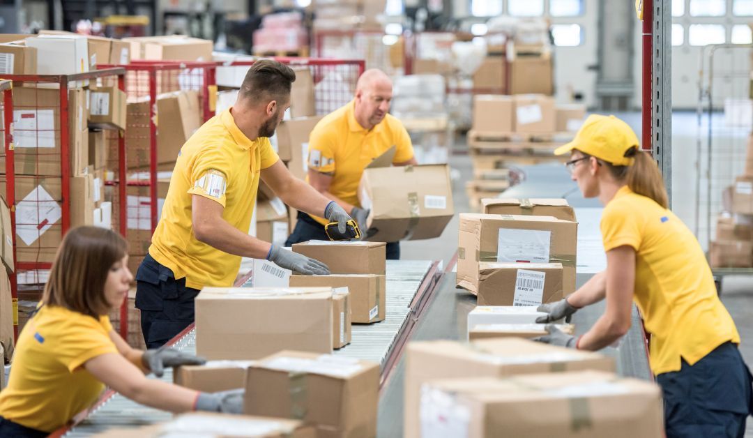 Warehouse workers stand near a conveyor belt and inspect packages as they go by.