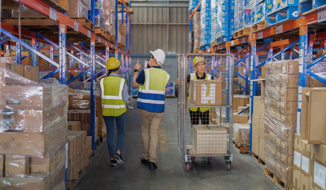 3 warehouse workers with safety hats and vests are walking down a warehouse aisle. A man and a woman have their backs facing the viewer, while a man pushing a cart is coming towards where the photo was taken.
