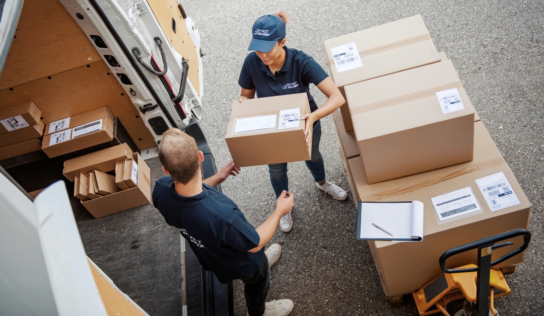 A woman hands a man a box from a stack of boxes, as they load a delivery van together.
