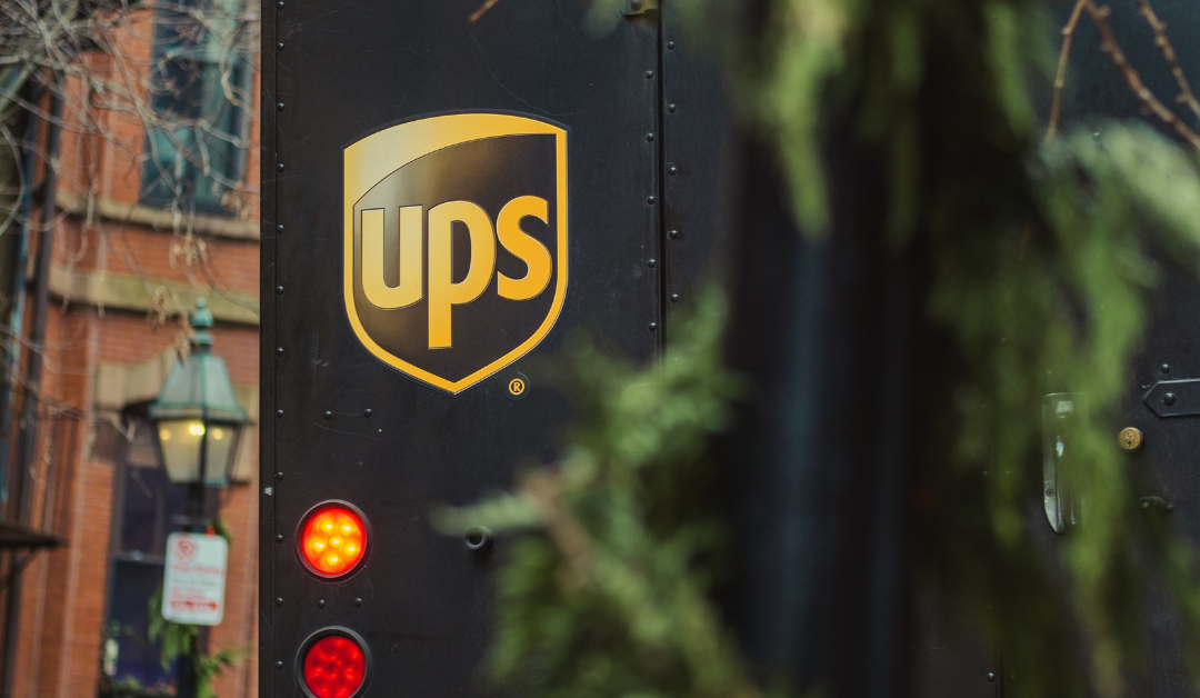 The backend of a UPS truck is hidden partially behind a tree with the logo visible.