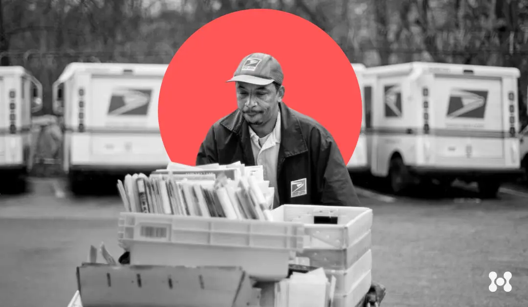 A USPS delivery man is shown wheeling a large bin of mail in a USPS truck parking lot.